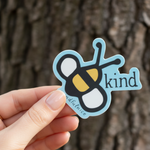 Hand holding Be Kind sticker in front of a tree