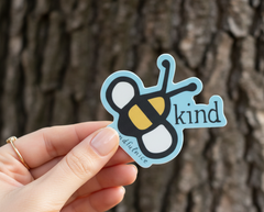 Hand holding Be Kind sticker in front of a tree