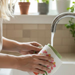 Person washing a floral towel under running water in a kitchen sink with plants in the background.